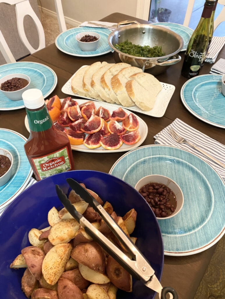 dinner table set with sliced oranges, steamed kale, sourdough bread, roasted potatoes, and black beans