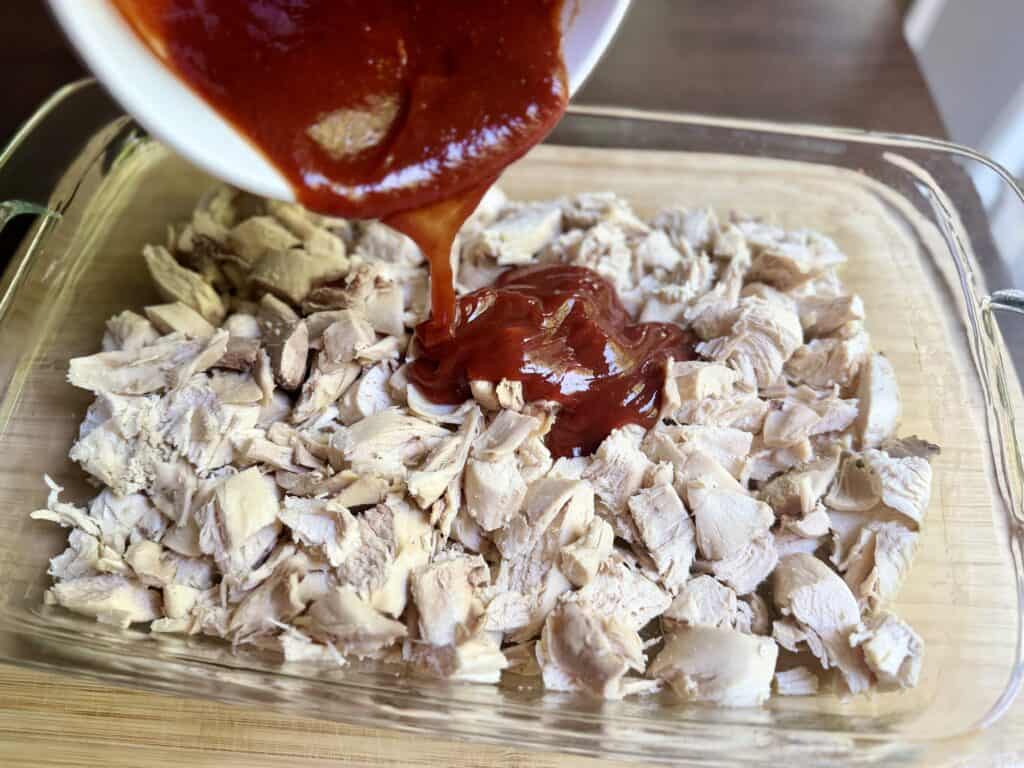 Aimee from Homemaker on a Budget pours BBQ sauce onto cooked chicken for a Hawaiian Chicken Bake casserole. 